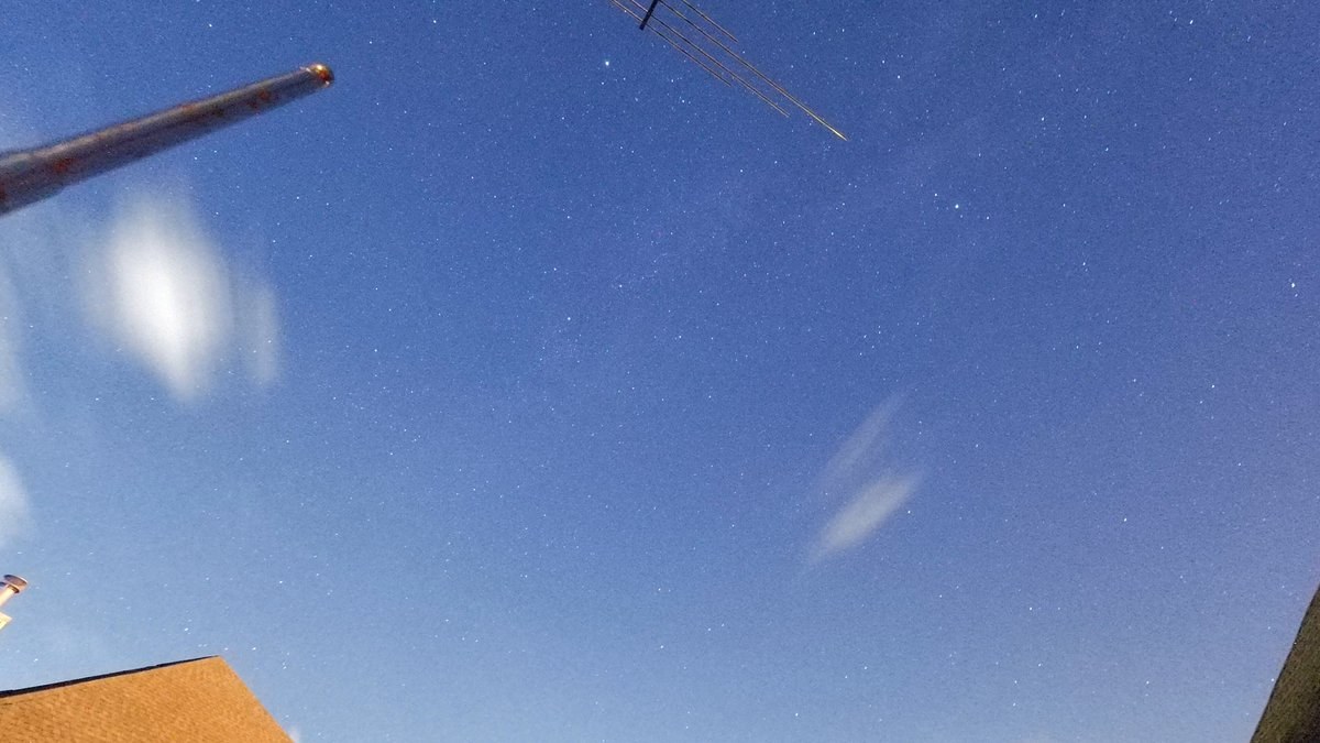 Night sky photo showing a faint meteor streak near a ham radio antenna mast, with a rooftop in the foreground