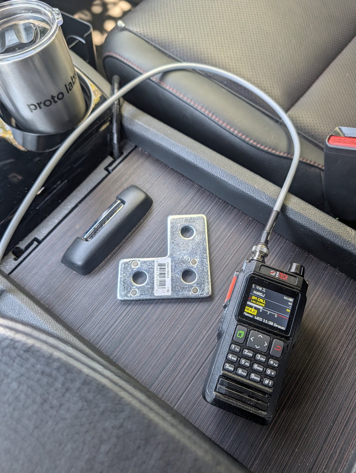 BTECH UV-PRO handheld radio and mounting hardware in a car center console next to a travel coffee tumbler