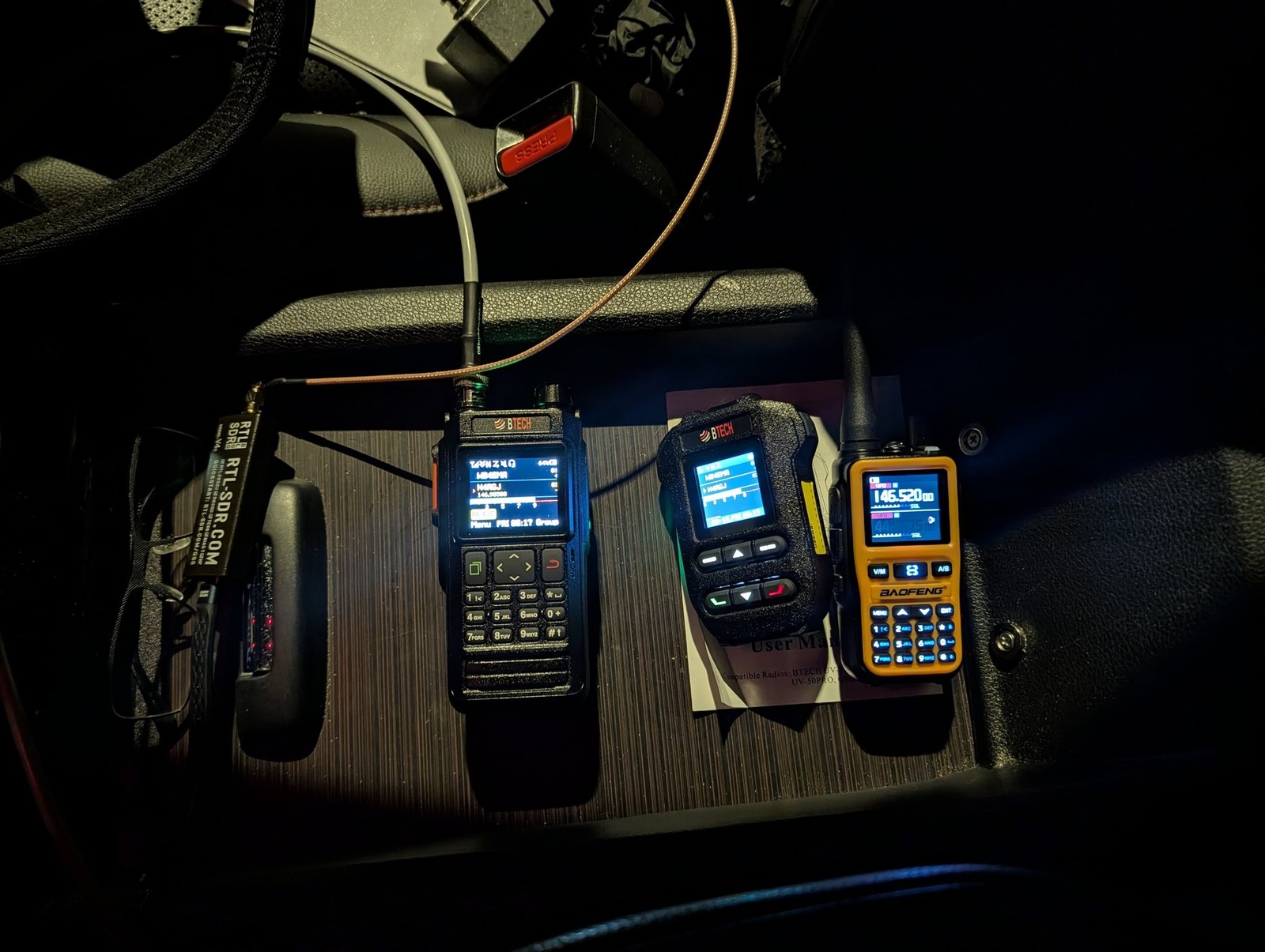Two BTECH handheld radios, a Baofeng handheld, and an RTL-SDR dongle in a car center console at night, screens lit showing a scan display and callsign N4RGJ